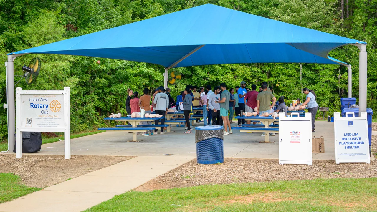 A free CPR Works community training event under a park picnic shelter, with learners practicing at tables and a Rotary sign out front