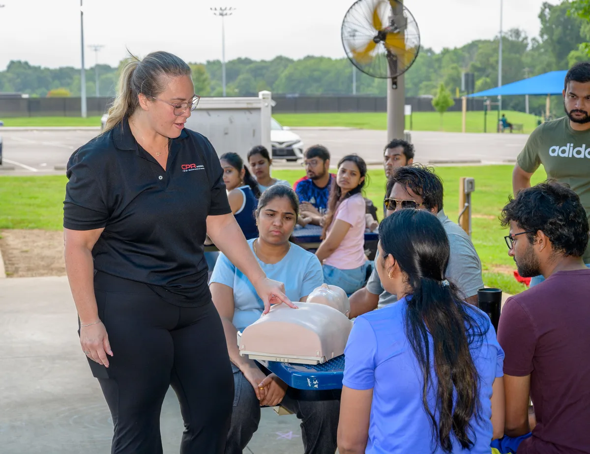 A CPR Works instructor addressing a seated group of learners with a hand resting on an adult training manikin