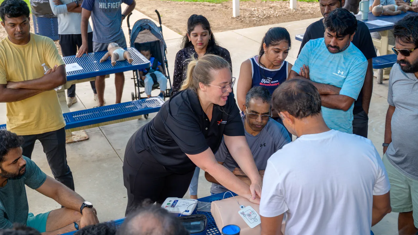A CPR Works instructor demonstrates compressions on a manikin as community members gather around picnic tables in the park
