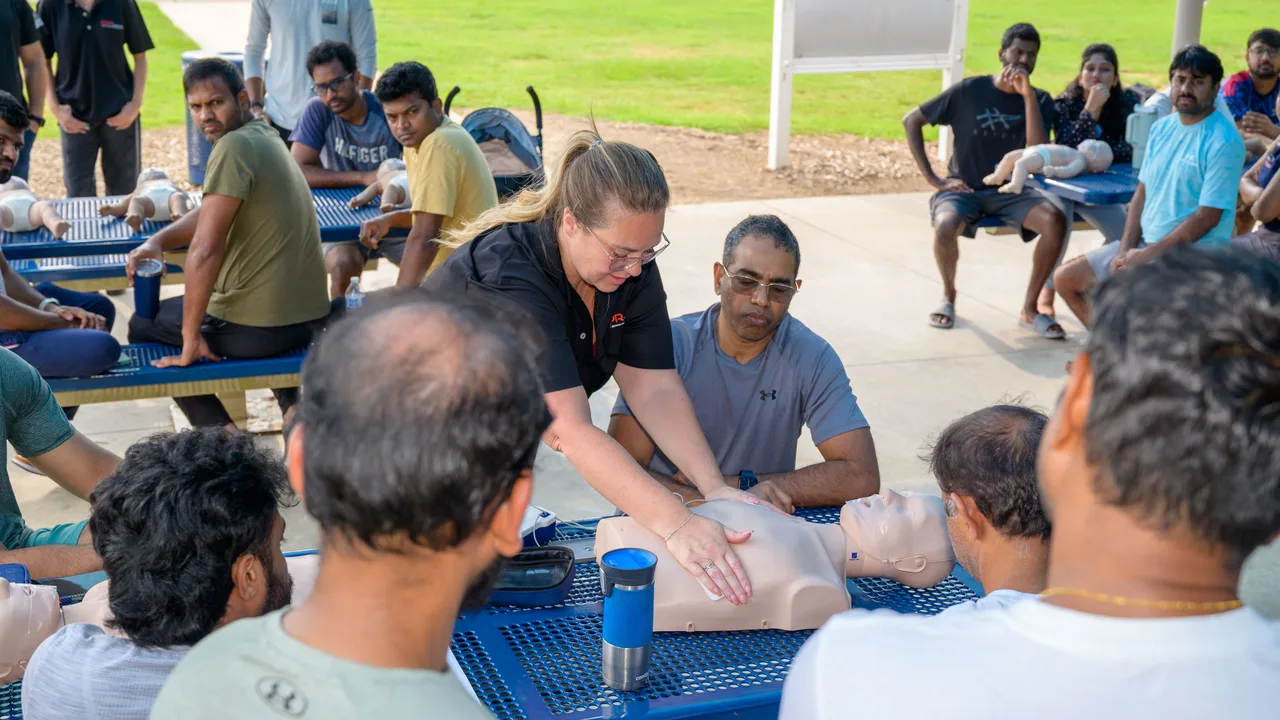 A CPR Works instructor shows hand placement on an adult training manikin to students seated around an outdoor table