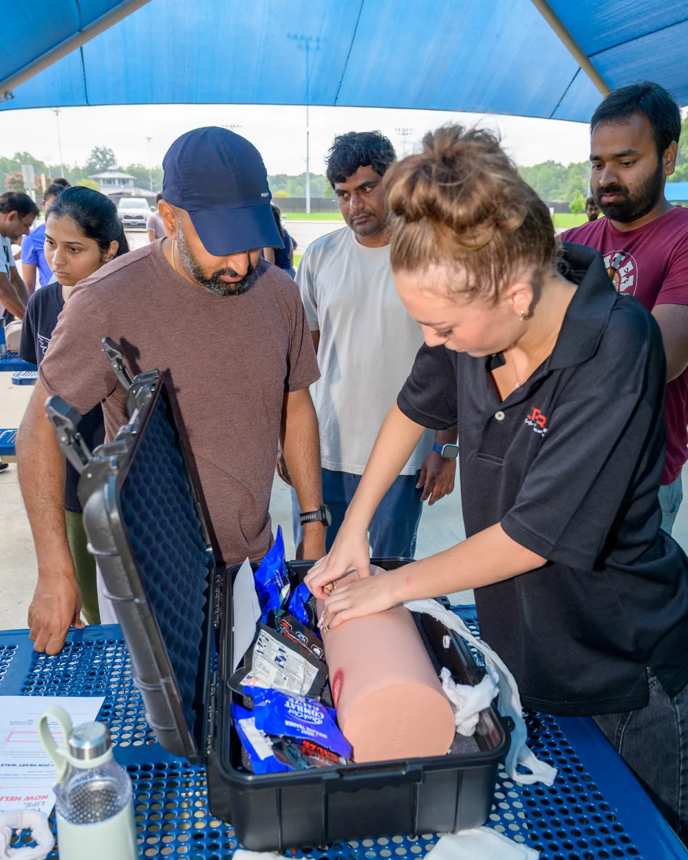A CPR Works instructor packs a wound on a bleeding-control trainer from an open Stop the Bleed kit while community members watch at an outdoor class