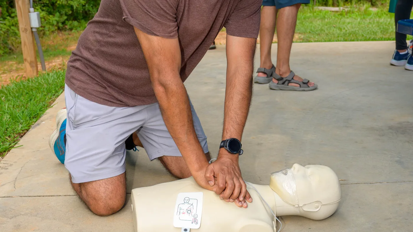 Un estudiante arrodillado al costado de un maniquí adulto comprime el centro del pecho con los dedos entrelazados, junto a un AED de entrenamiento abierto
