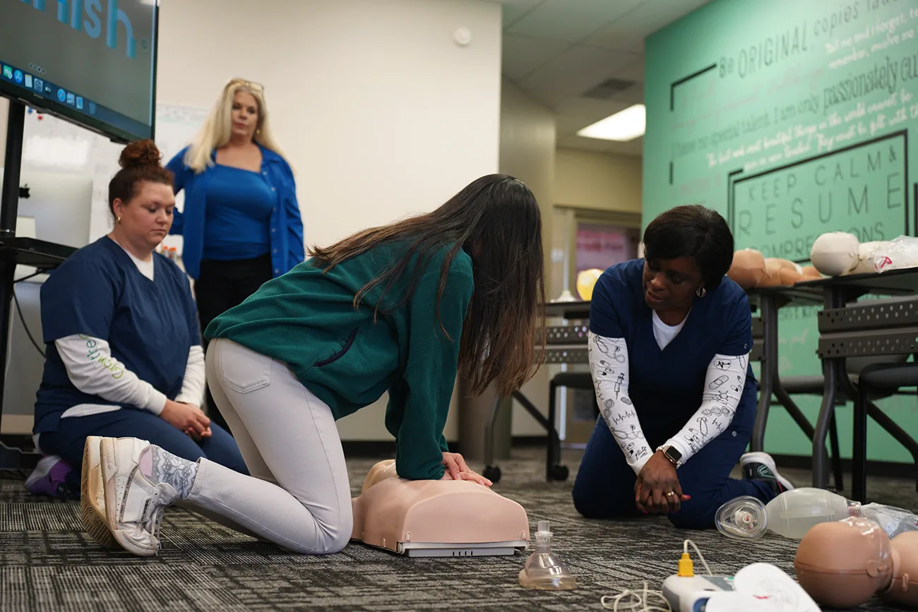 A student kneels to practice chest compressions on a training manikin while two CPR Works instructors in scrubs watch and coach during a classroom course