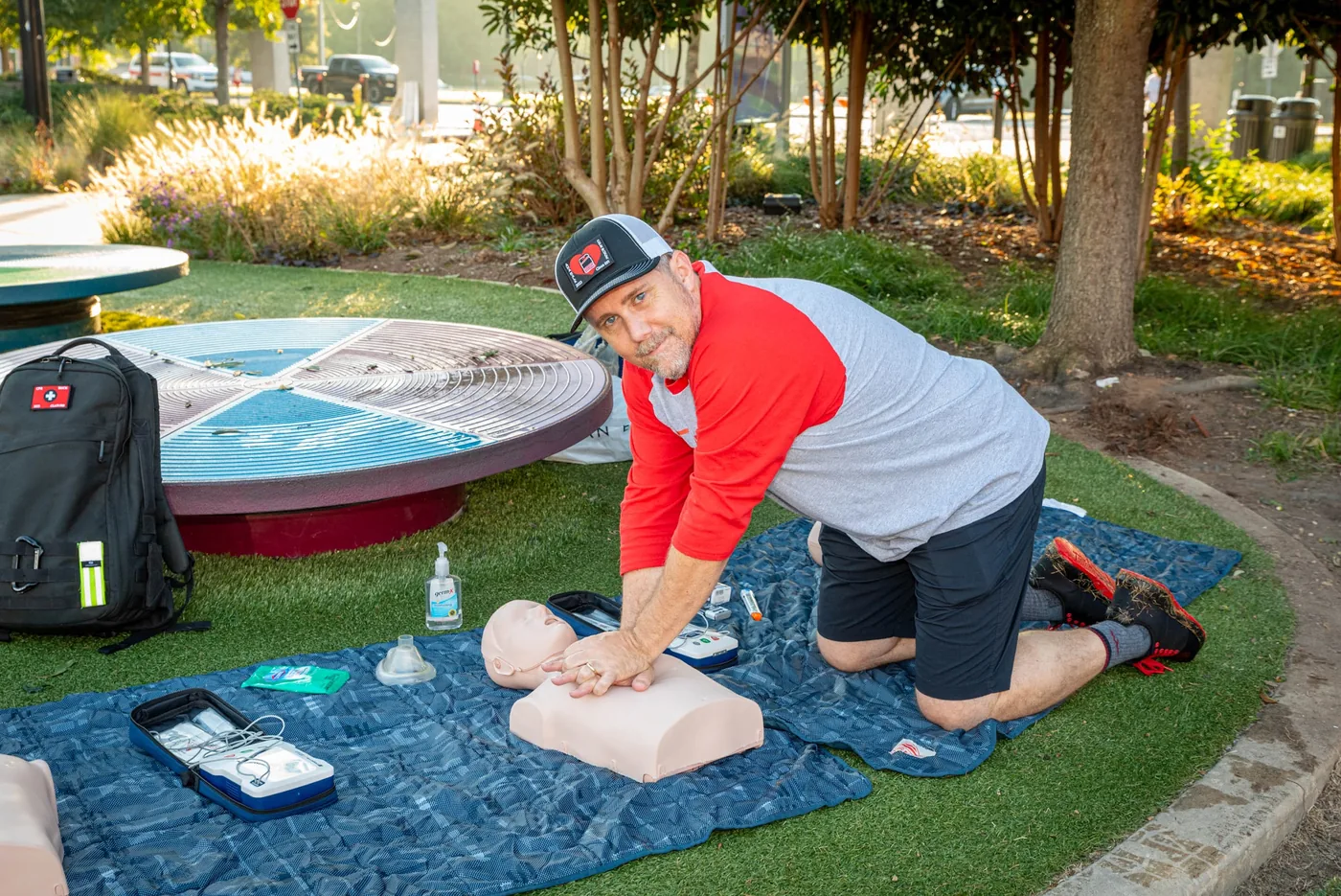 A CPR Works instructor kneels on a blanket in a park to demonstrate chest compressions on a training manikin, with an AED trainer and supplies beside him