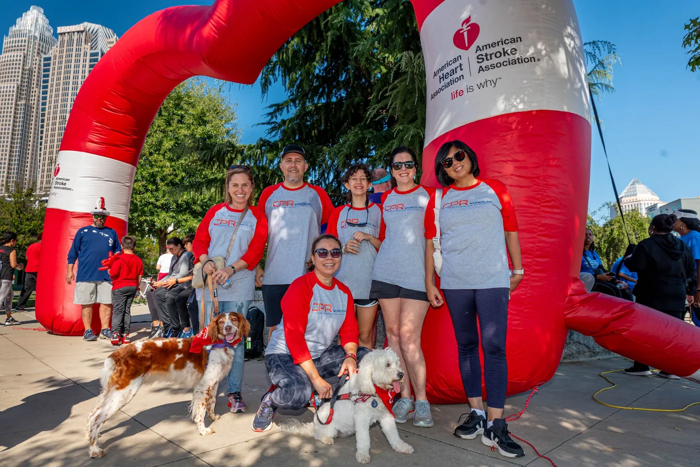 The CPR Works team in matching CPR Works shirts with two dogs under the American Heart Association arch at the Charlotte Heart Walk, with the uptown skyline behind them