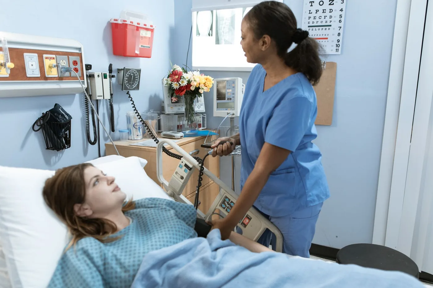 A nurse checks the bedside monitor while talking with a patient lying in a hospital bed