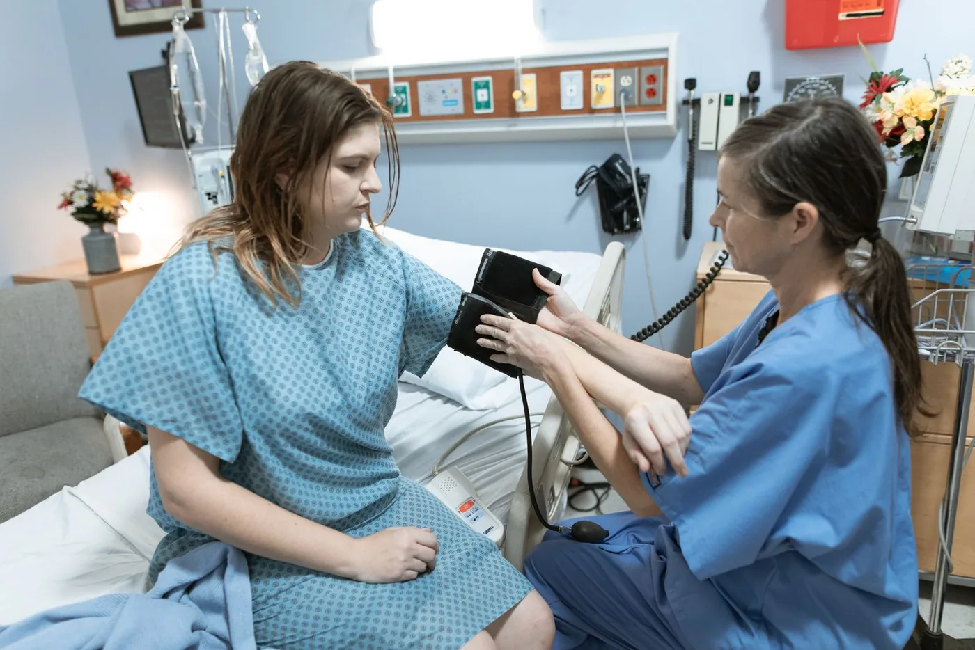 A nurse in blue scrubs takes the blood pressure of a patient sitting on the edge of a hospital bed